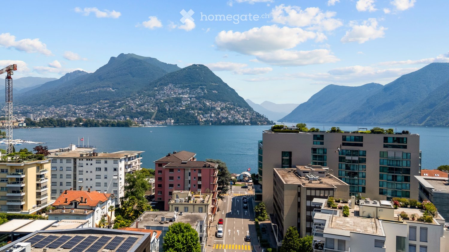 A panoramic view of a cityscape with a lake and mountains in the background, featuring modern and traditional buildings, a road, and a construction crane.
