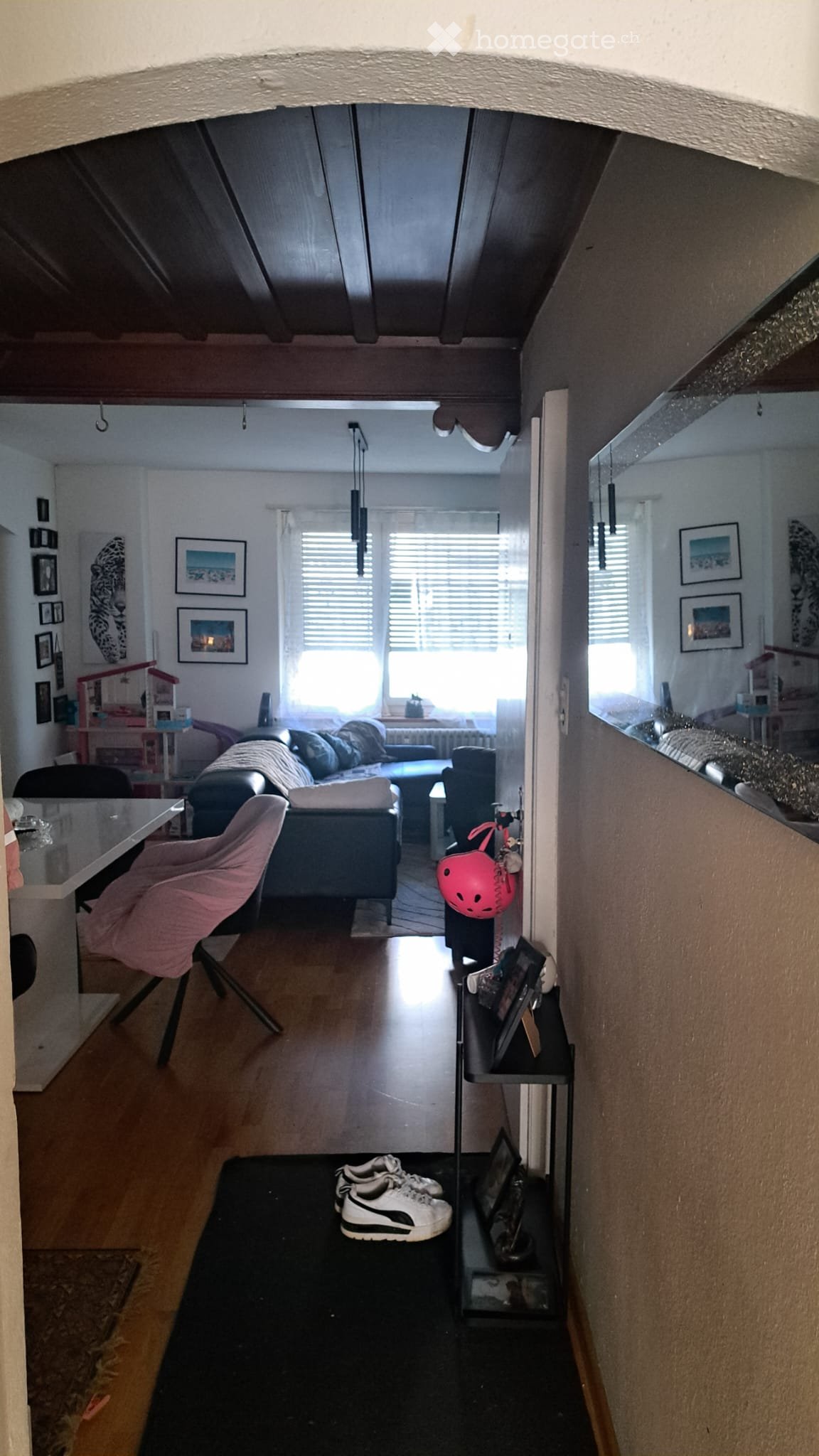 A modern living room with a sofa, chairs, a glass table, a mirror, picture frames, and a shelf with a helmet and shoes.