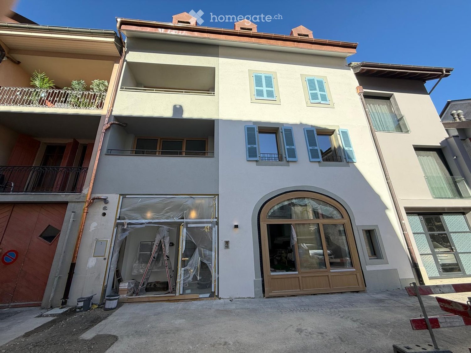 Two-story residential building with light-colored facade, multiple windows with shutters, balconies, and a covered entrance area under construction.