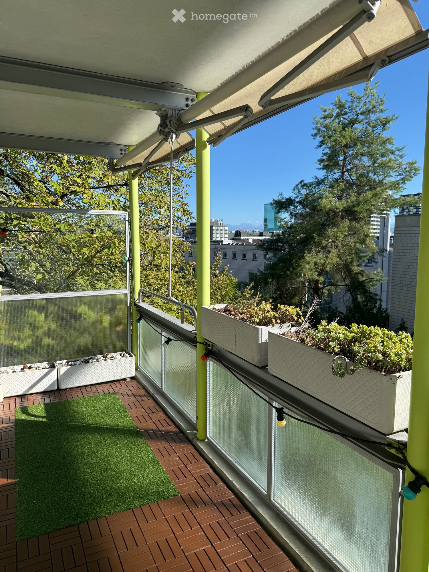 Balcony with wooden decking, green artificial grass, planters with plants, and a city view.