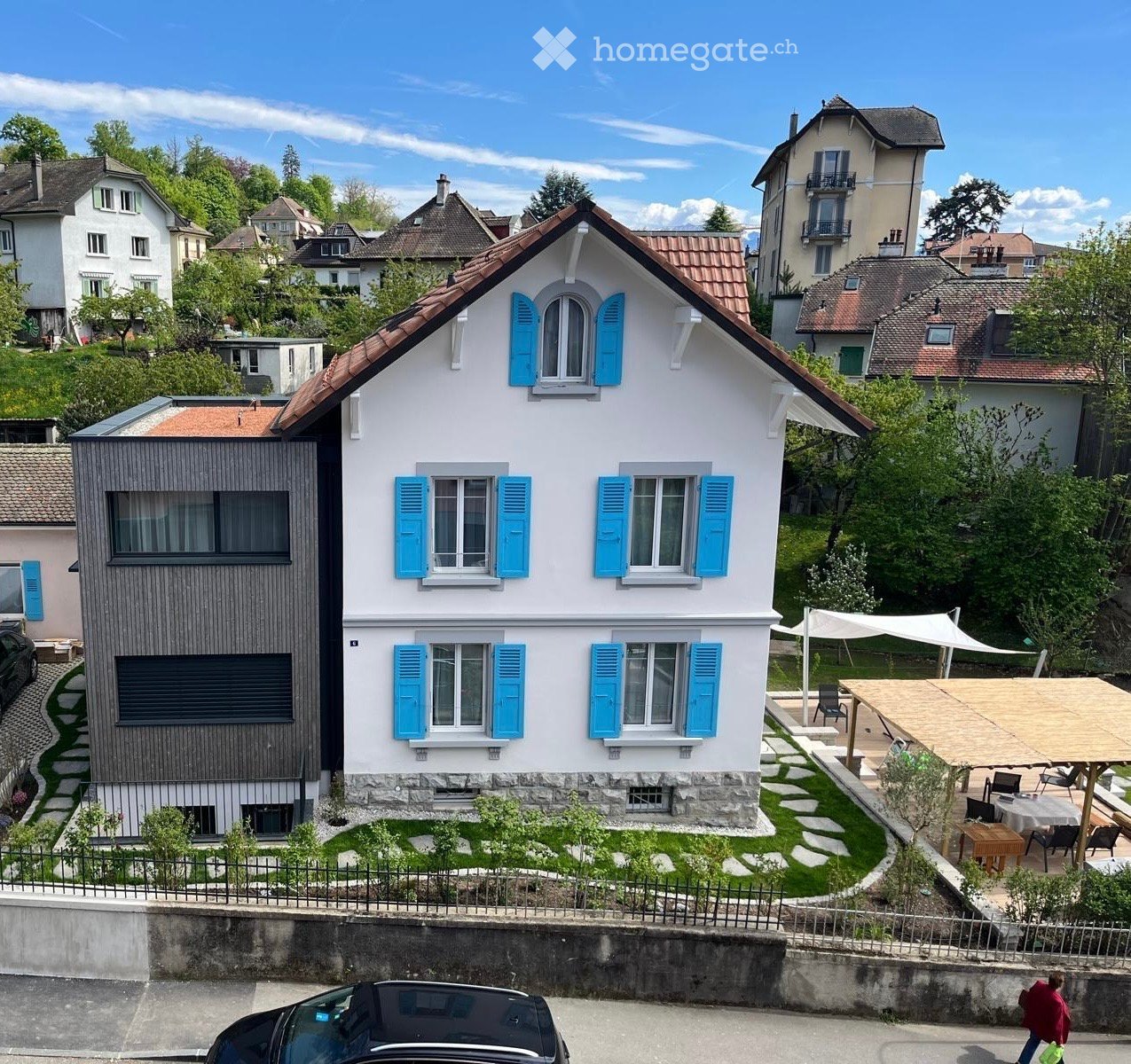 Two-story house with white walls, blue shutters, and a red-tiled roof. The house features a modern extension with dark wooden panels and large windows. The front yard includes a small garden with stepping stones and a covered outdoor seating area with tab