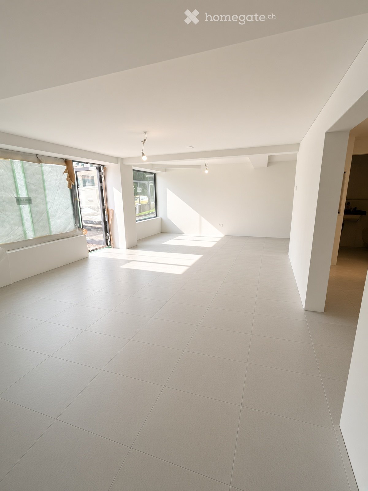 Empty room with white tiled flooring, white walls, and large windows allowing natural light. No furniture or equipment is present.