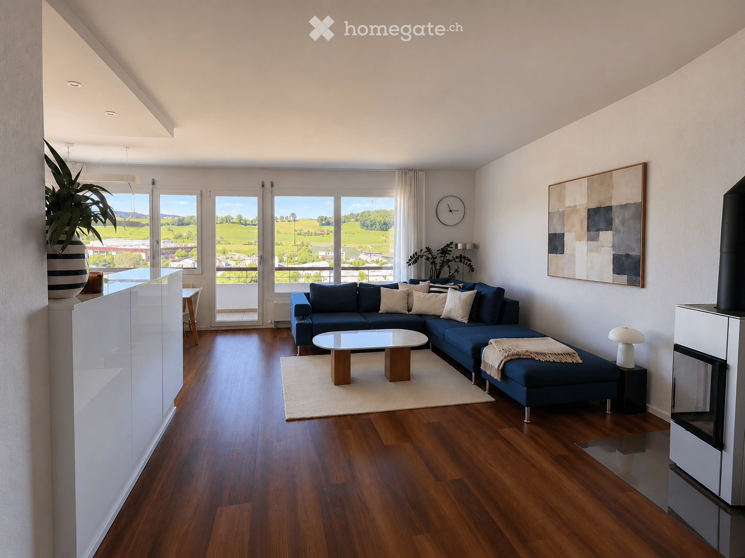 Modern living room with wooden flooring, large windows offering a scenic view, a blue sectional sofa, a glass coffee table, a small dining table with chairs, a lamp, a clock, a painting, and a fireplace.