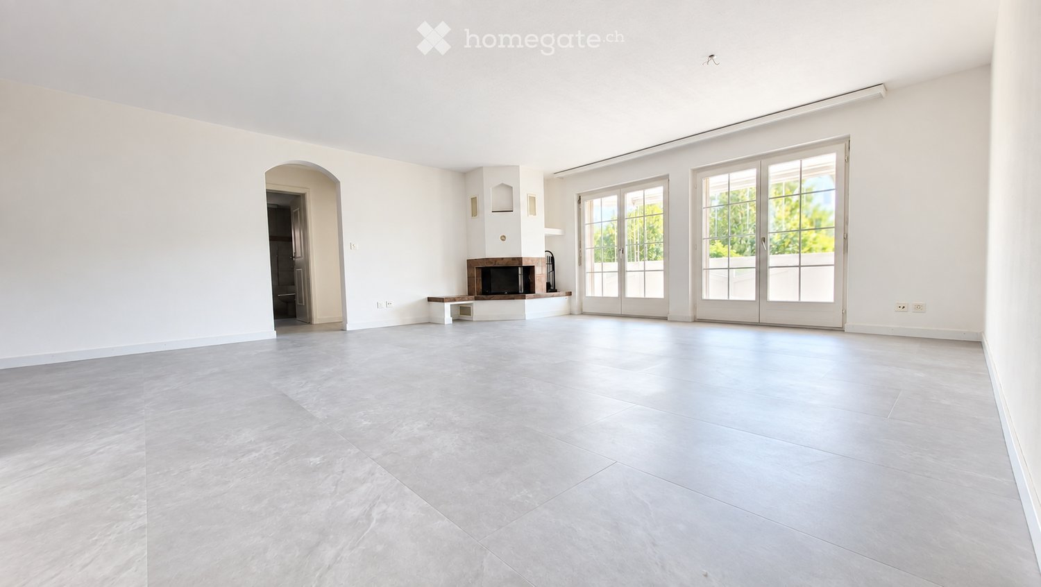 Empty living room with white walls, grey tiled floor, large glass doors, and a fireplace area.