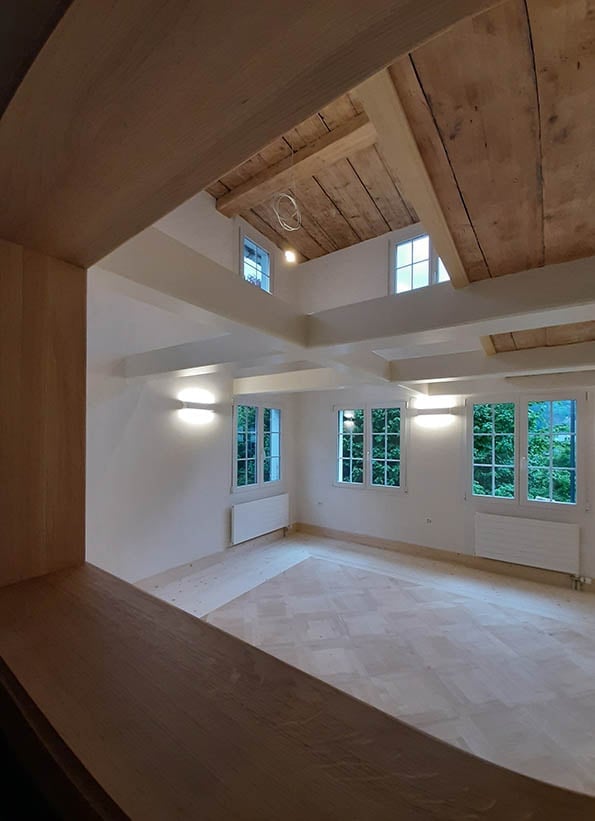 Empty room with wooden ceiling beams, large windows, and light-colored flooring.
