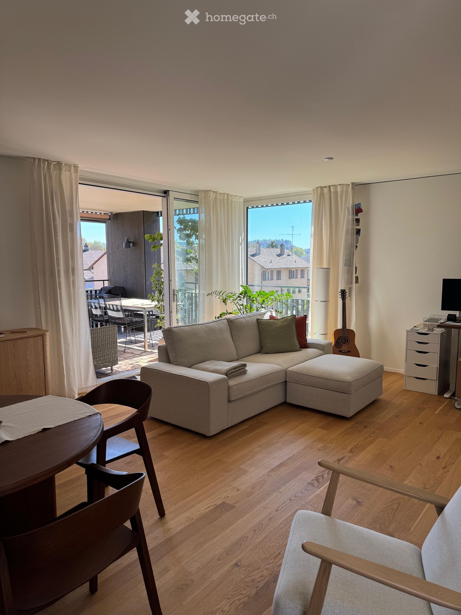 Modern living room with wooden flooring, large windows, and a balcony. Features a sectional sofa, wooden dining table, chairs, and a guitar.