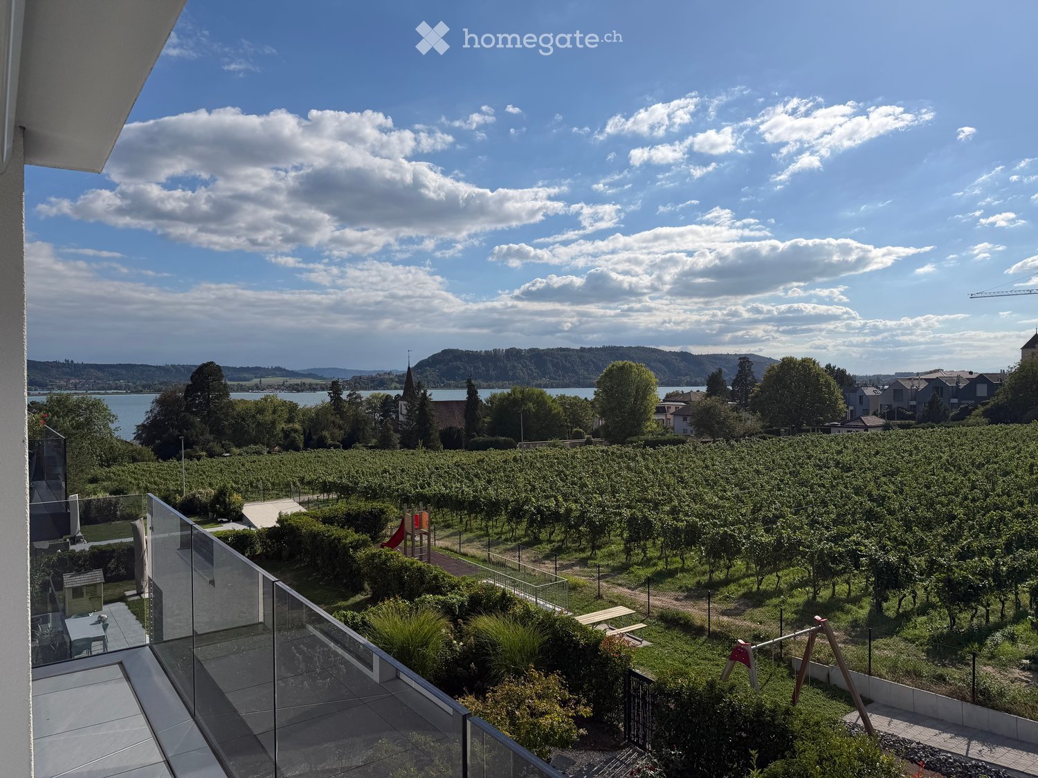 Balcony overlooking a vineyard, lake, and distant hills under a partly cloudy sky.