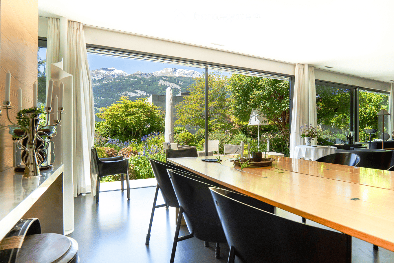 Bright room with large windows offering a scenic mountain view, furnished with a long wooden table, chairs, a decorative candle holder, and a vase on a side table.