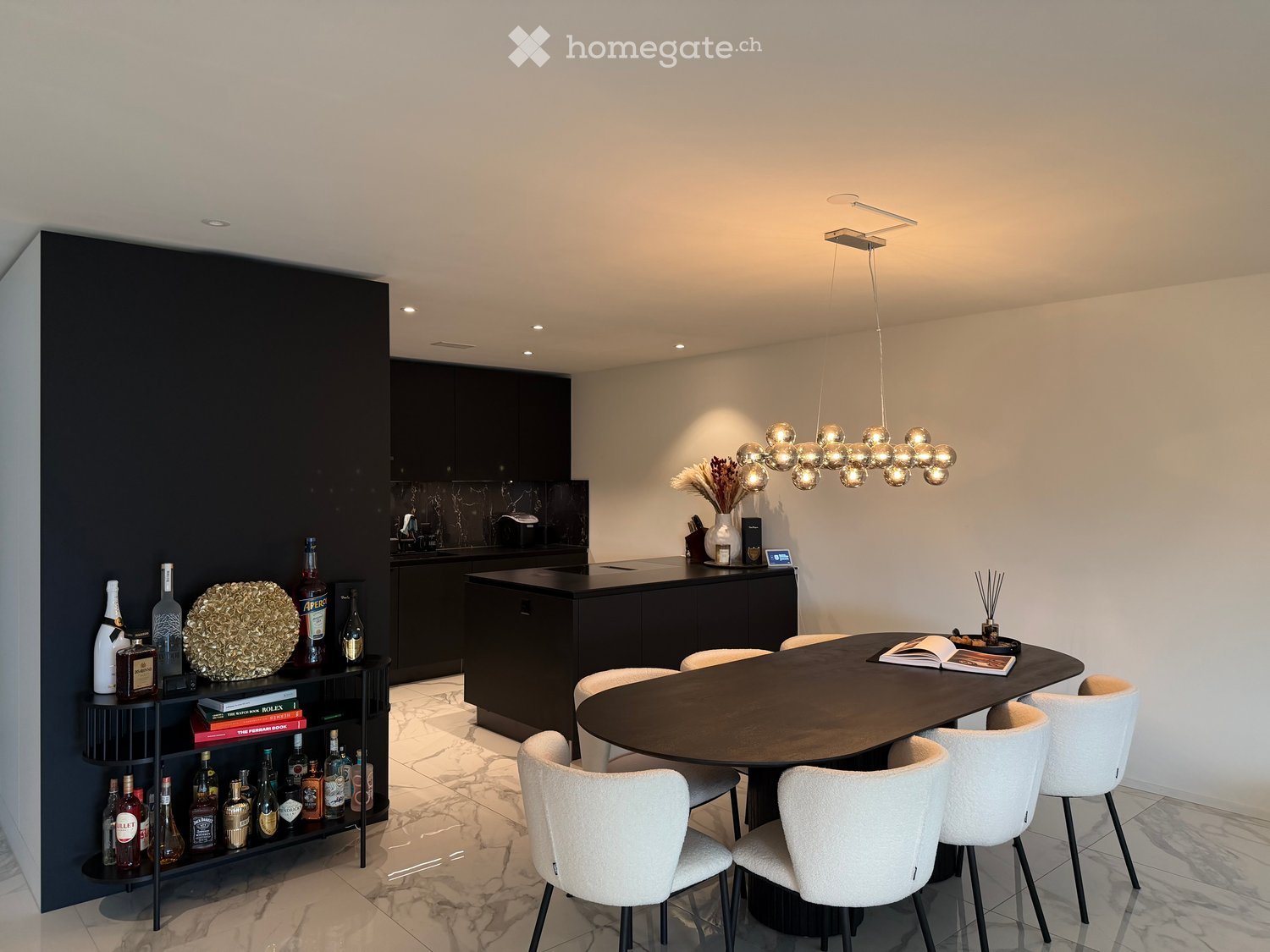 Modern kitchen and dining area with a round dining table, white chairs, a dark cabinet, and a shelf with bottles and decorative items.