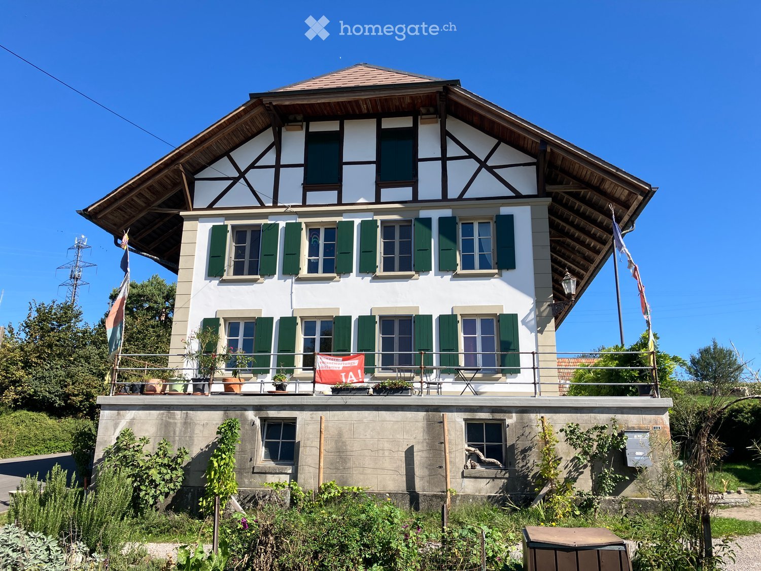 Two-story house with white walls, green shutters, and a wooden roof. Features a raised ground floor, balcony with potted plants, and a garden area.