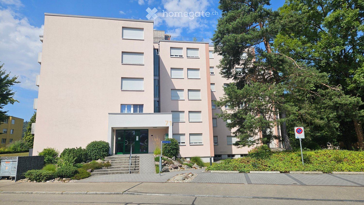 A modern, multi-story residential building with a light pink facade, rectangular windows, and a small entrance area with steps and greenery.