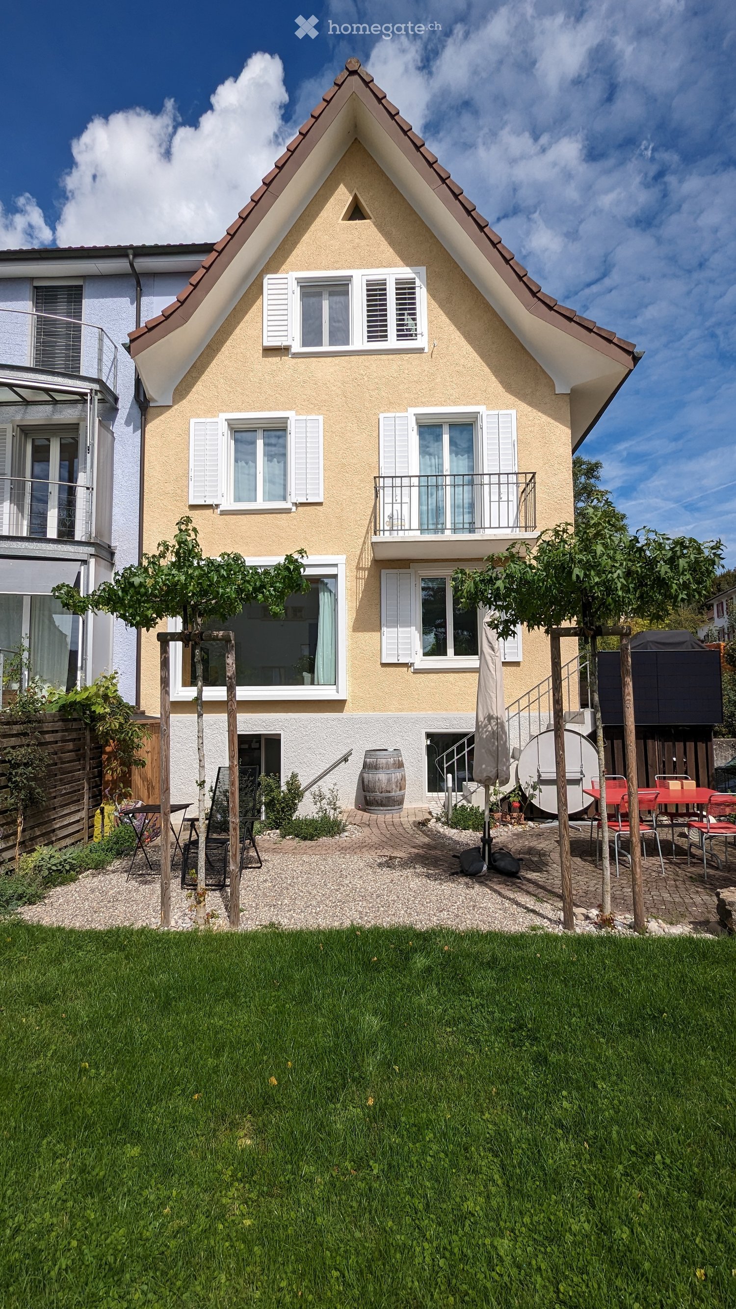Two-story house with a modern design, featuring a triangular roof, multiple windows with shutters, a balcony, and a well-maintained garden with seating area.
