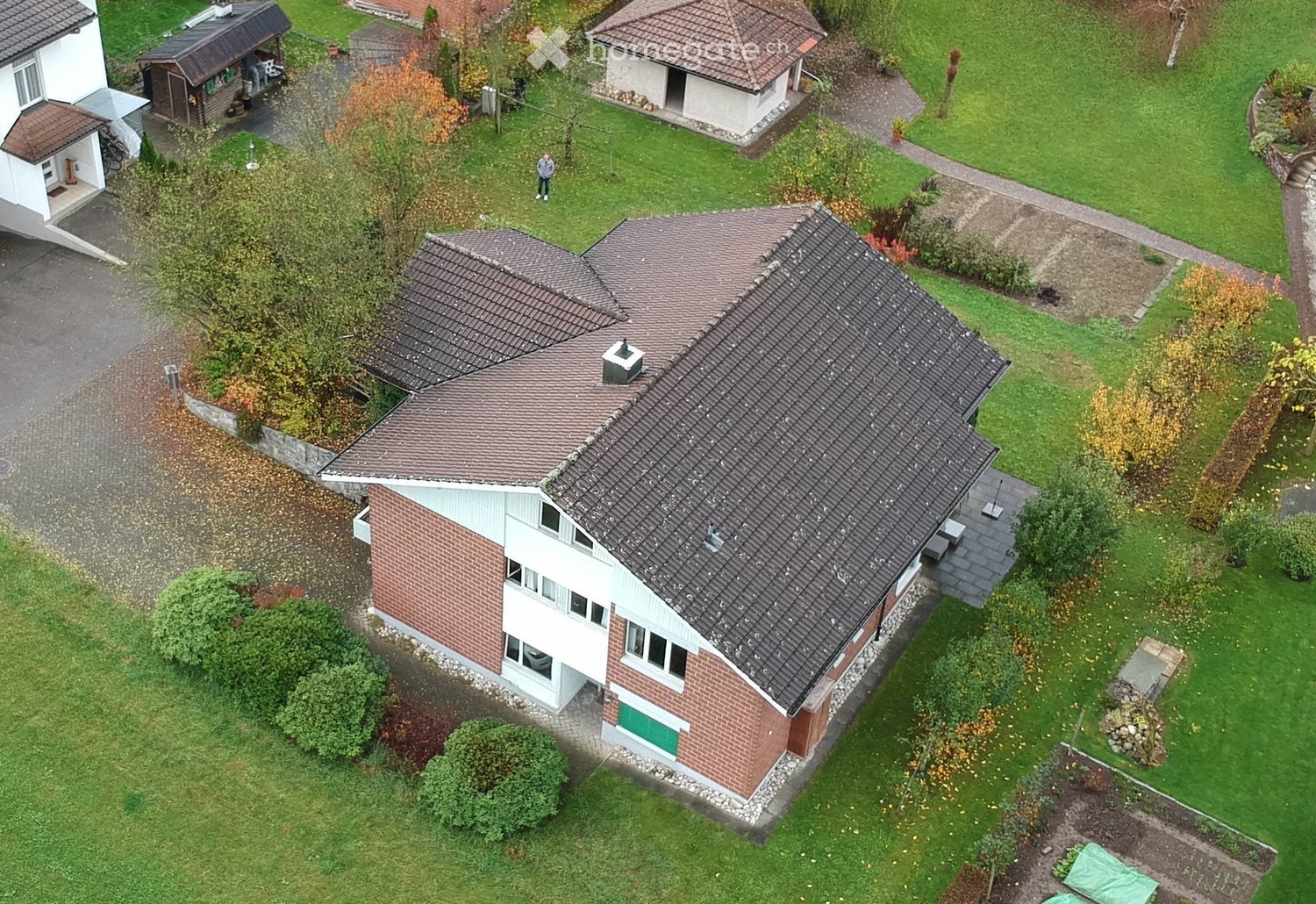 Aerial view of a two-story house with a brown tiled roof, surrounded by a well-maintained garden with trees, shrubs, and pathways.