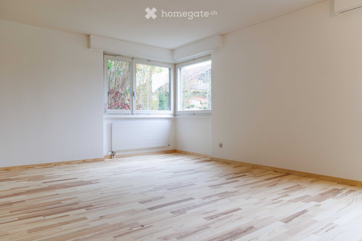 Empty room with light wooden flooring, white walls, and large windows offering a view of greenery outside. A radiator is visible on one wall.