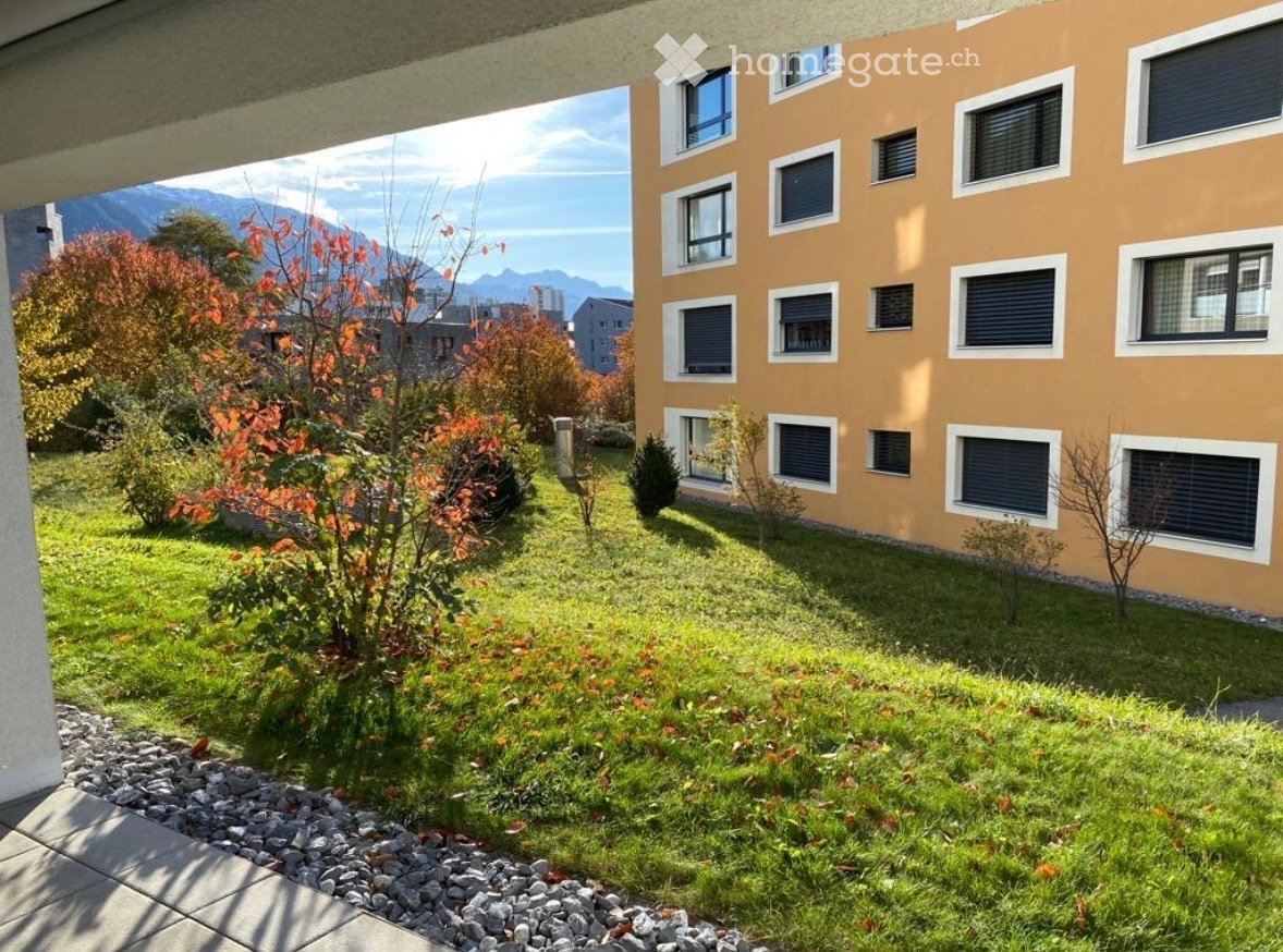 Well-maintained garden area with green grass, small shrubs, and a view of mountains in the background. The building has a light brown facade with multiple windows.