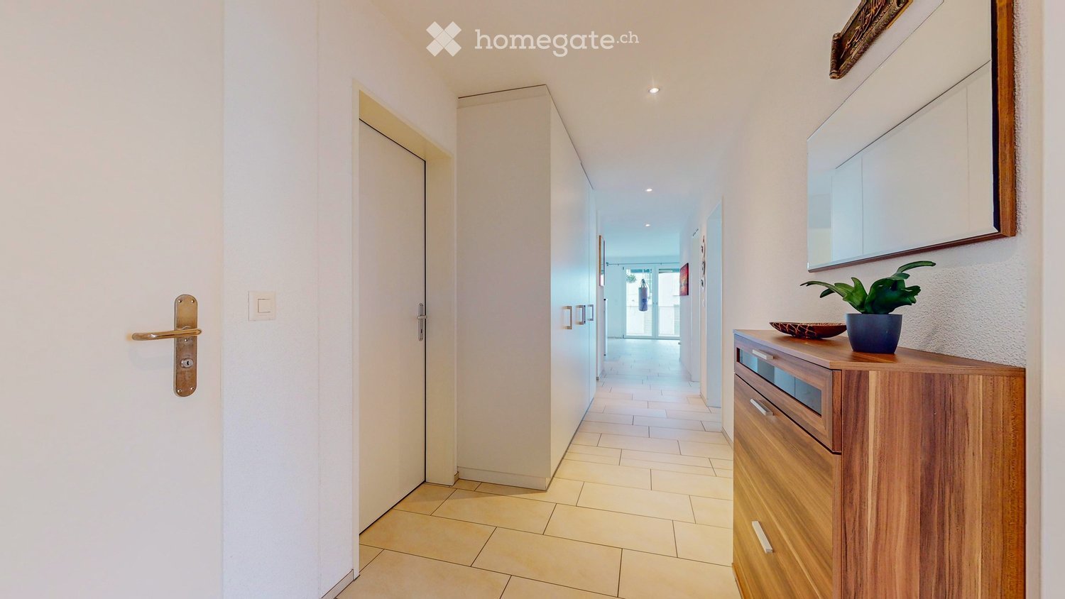 Bright hallway with tiled floor, white walls, and a small wooden console table with a potted plant and decorative bowl on top. A closed door with a brass handle is visible on the left.