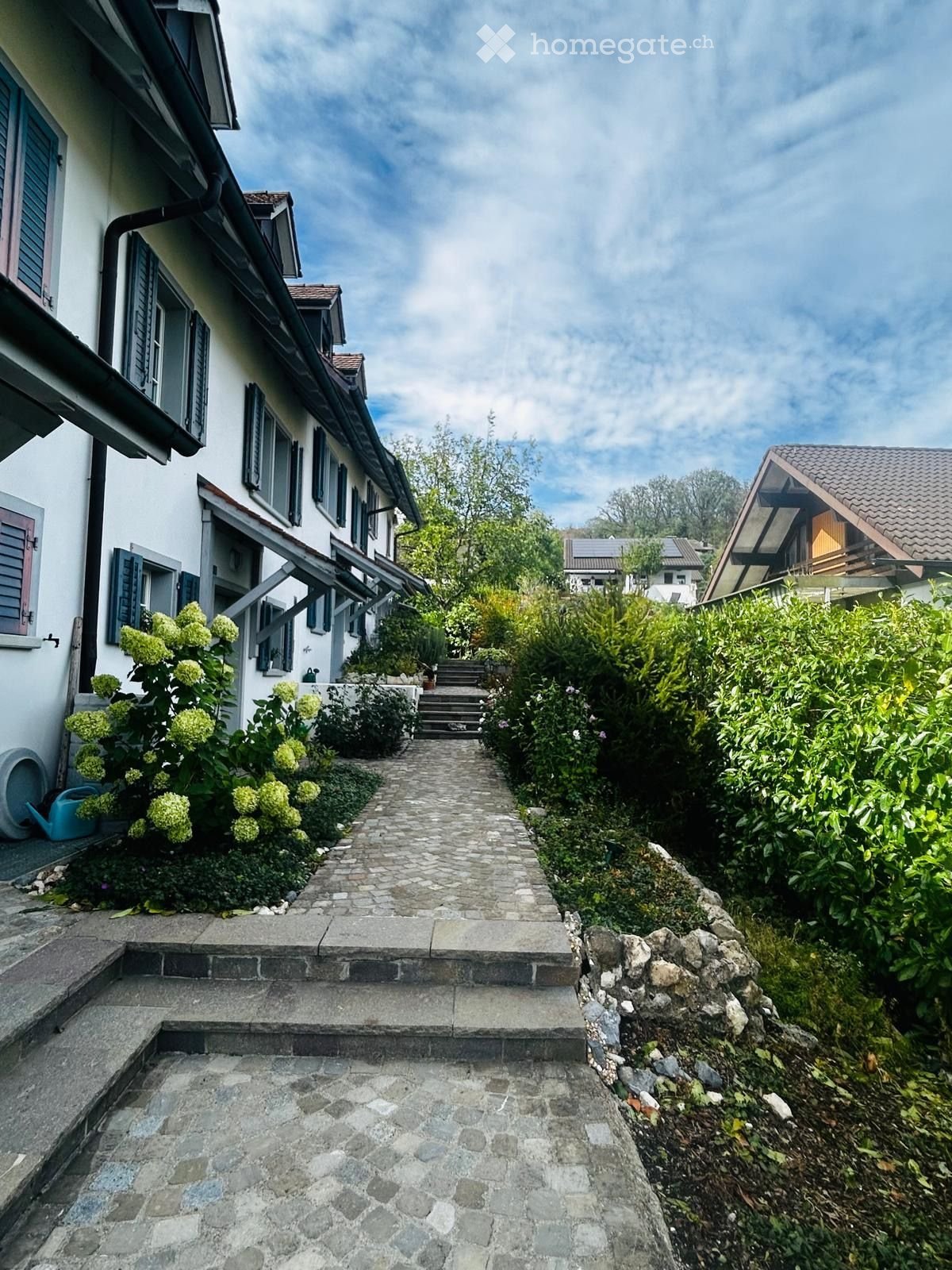 A stone pathway leads up to a house with white walls and dark shutters, surrounded by lush greenery and flowering bushes.