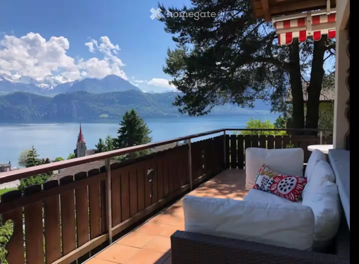 Balcony with wooden decking, white sofa, colorful pillow, overlooking a lake and mountains.