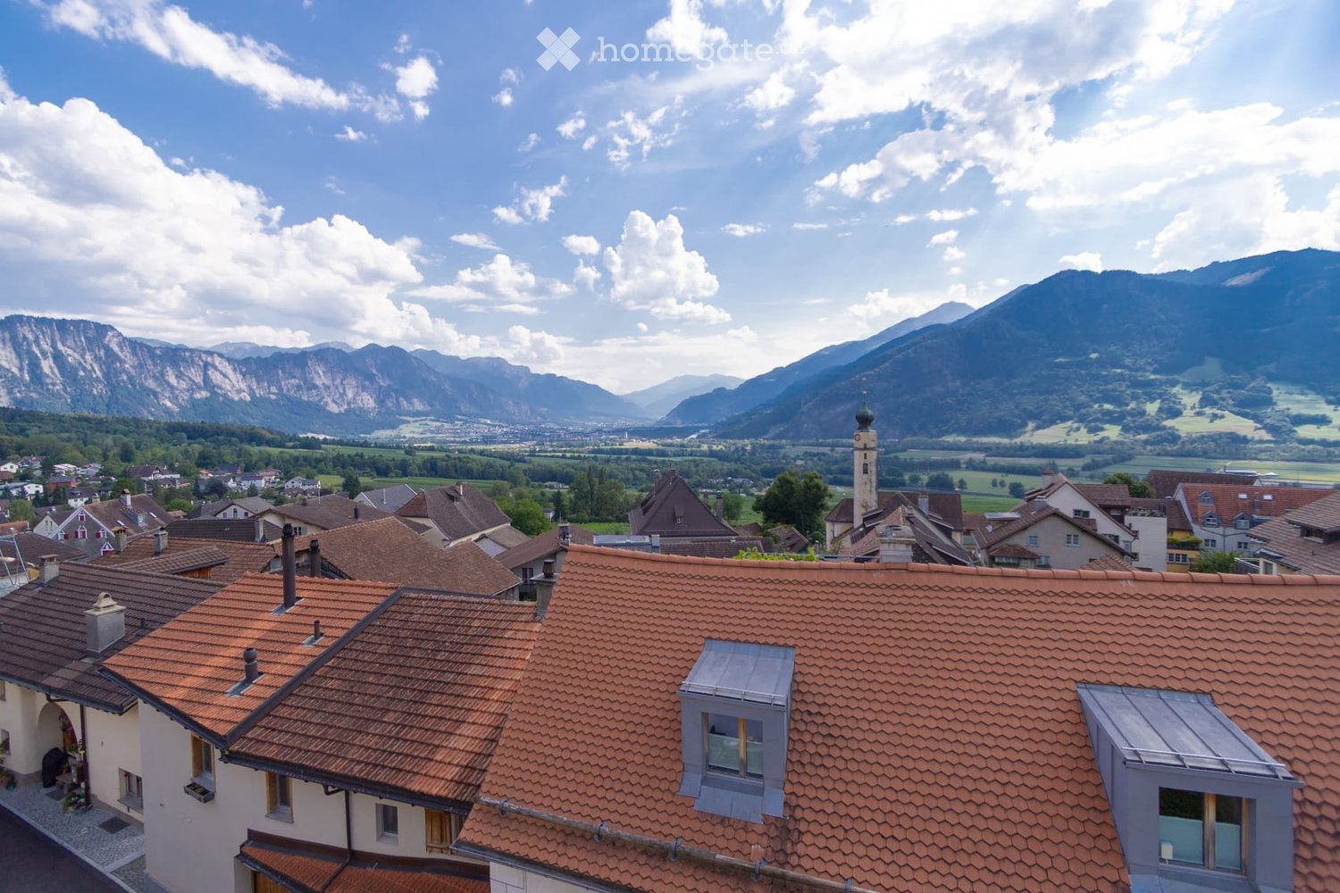 A panoramic view of a village with red-tiled roofs, surrounded by green fields and distant mountains under a partly cloudy sky.