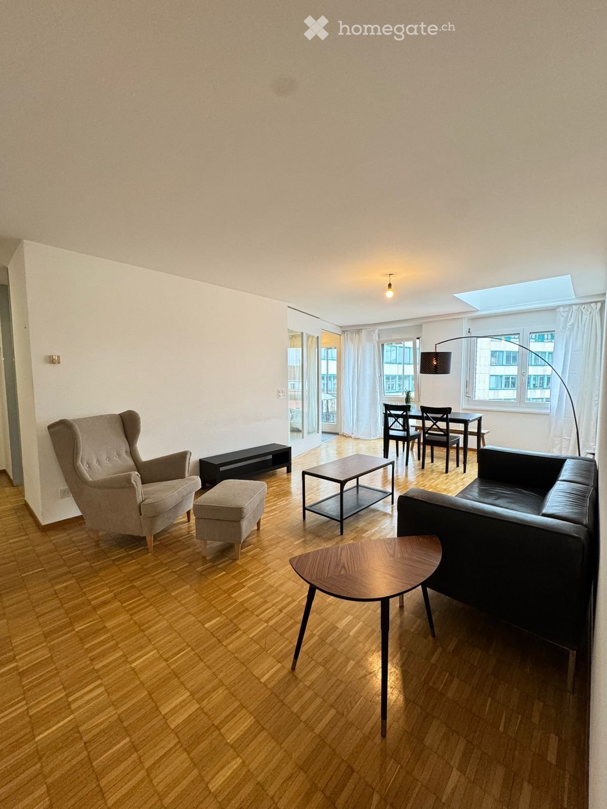 Modern living room with wooden flooring, a grey armchair, a black sofa, a small coffee table, a round wooden side table, a black bench, and a dining area with a table and chairs.