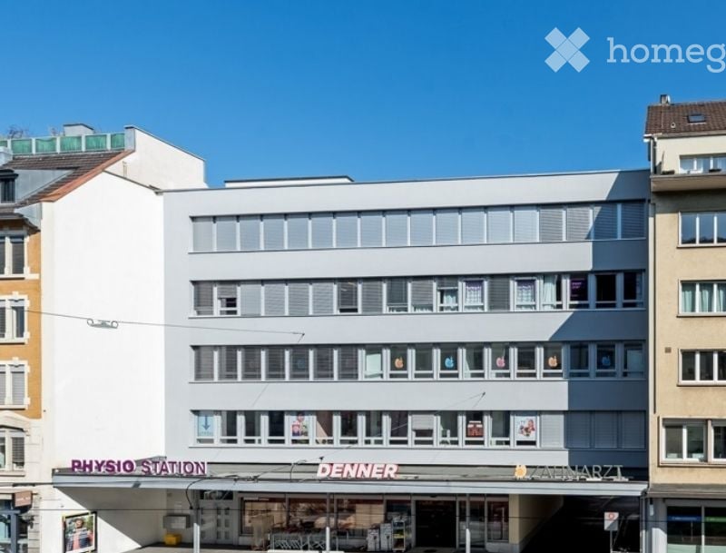 Modern multi-story building with a flat roof and large windows, featuring commercial signage for "Physio Station" and "Denner" on the ground floor.