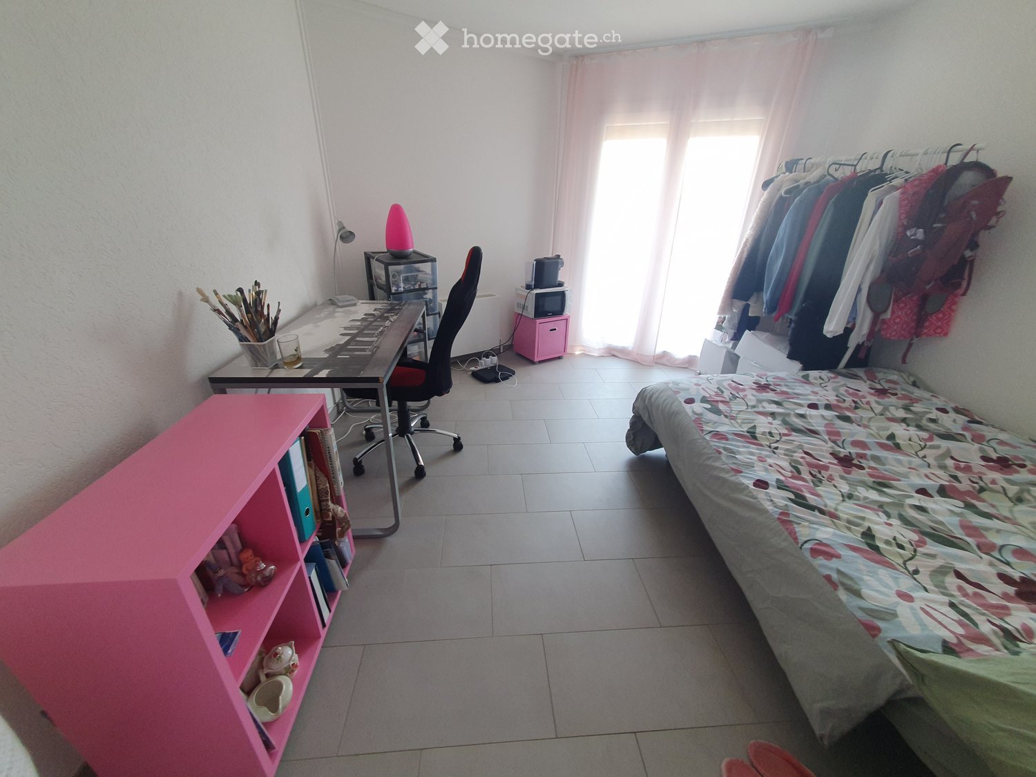 A small bedroom with a single bed, desk, chair, pink shelf, and a clothing rack. The room has tiled flooring and white walls.