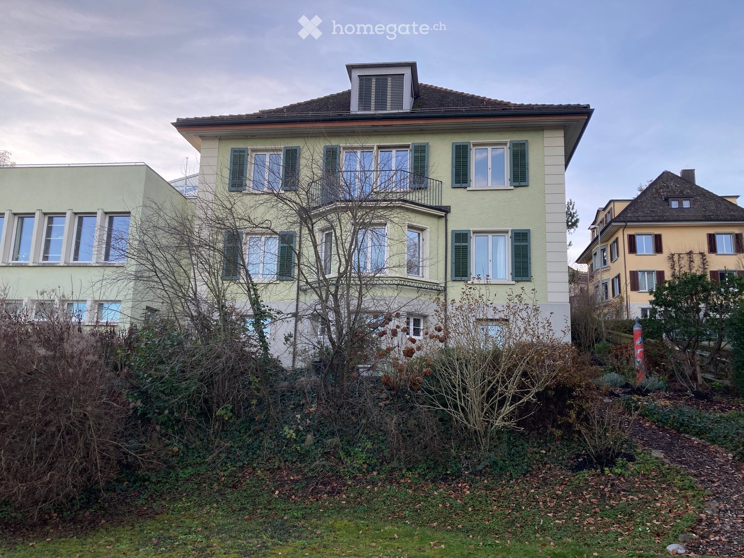 A multi-story residential house with light green walls, multiple windows with shutters, and a small balcony on the upper floor. The house is surrounded by a garden with various plants and trees.