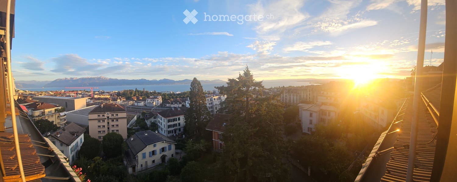 Balcony with a panoramic view of a cityscape at sunset, featuring buildings, trees, and distant mountains and water.