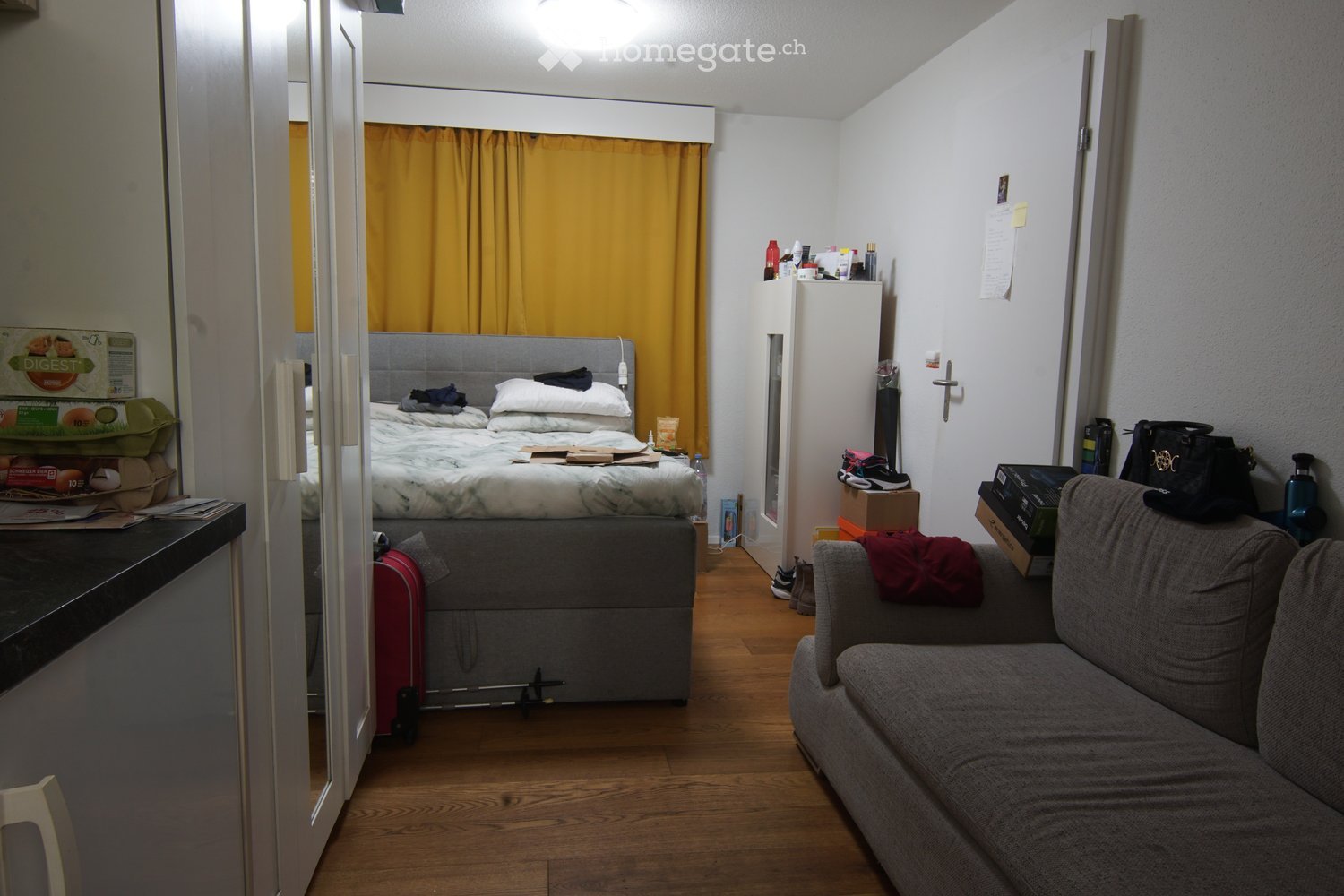 A small, furnished room with a bed, sofa, and a refrigerator. The room features wooden flooring, a grey sofa with a red blanket, a bed with pillows and a duvet, and various personal items including a suitcase, handbag, and laptop.