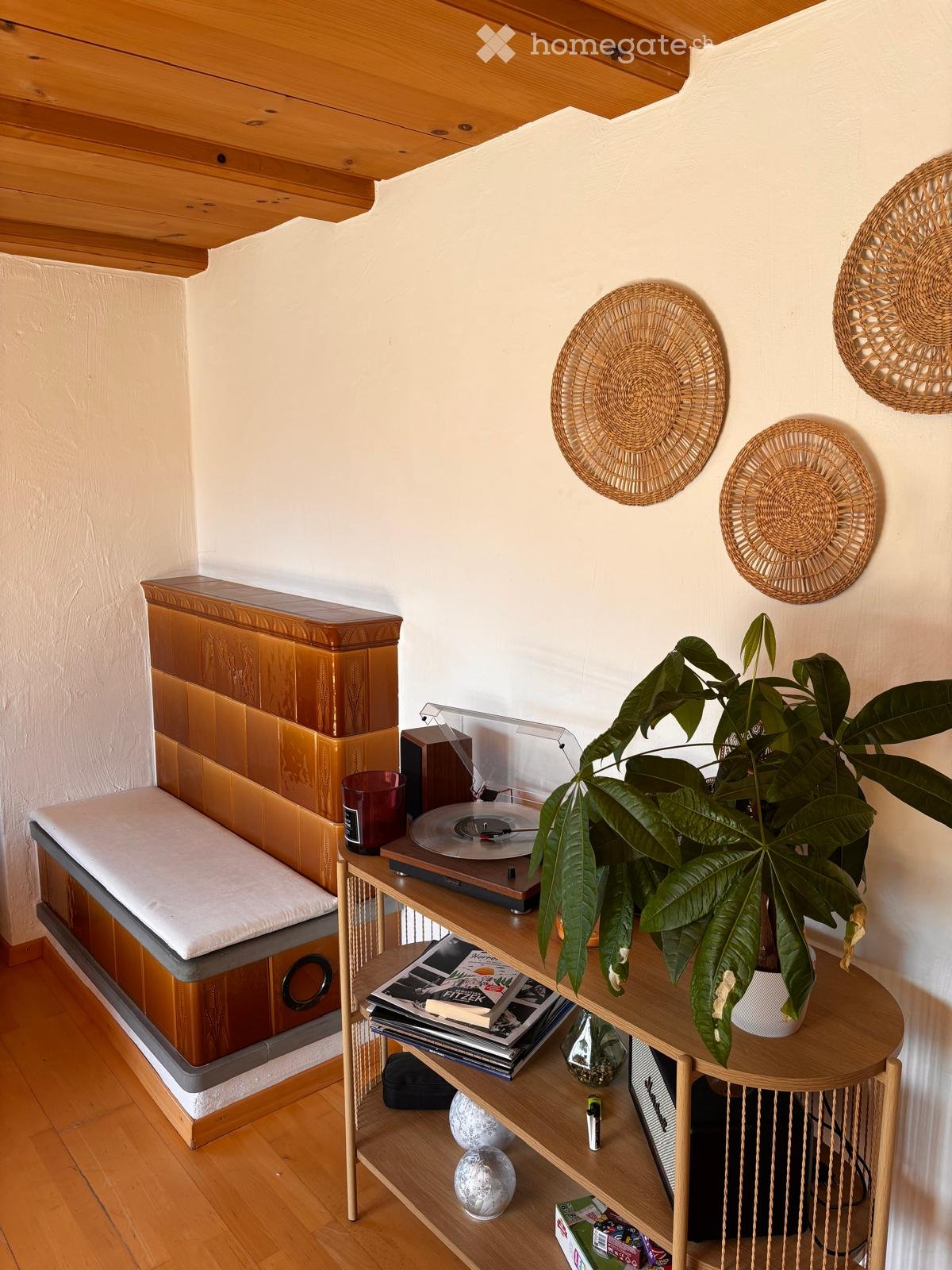 A cozy, well-furnished room with a wooden bed, a round wooden table holding a large leafy plant, a record player, and decorative wall hangings.