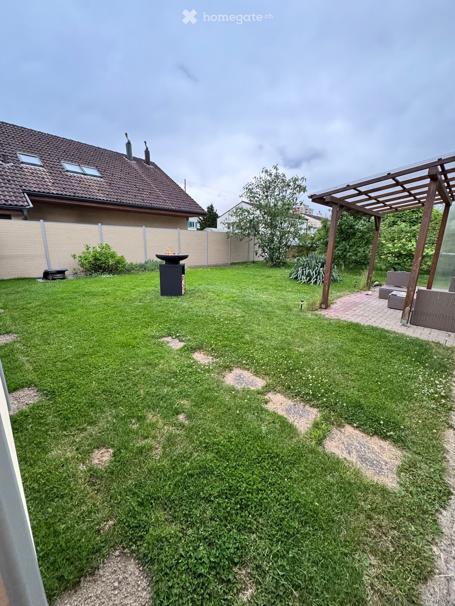 Well-maintained garden with a central fire pit, wooden pergola, and seating area, surrounded by a house and fence.