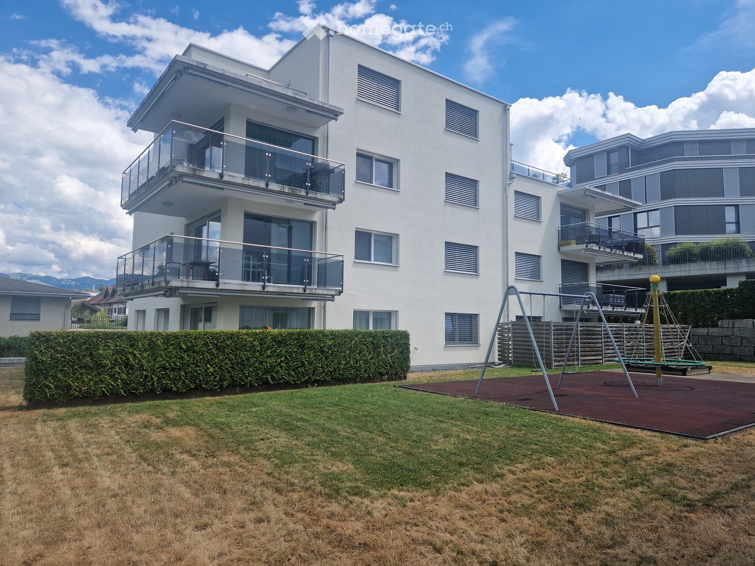 Modern two-story white building with large windows and balconies, surrounded by a well-maintained lawn and a playground area with swings and a climbing frame.