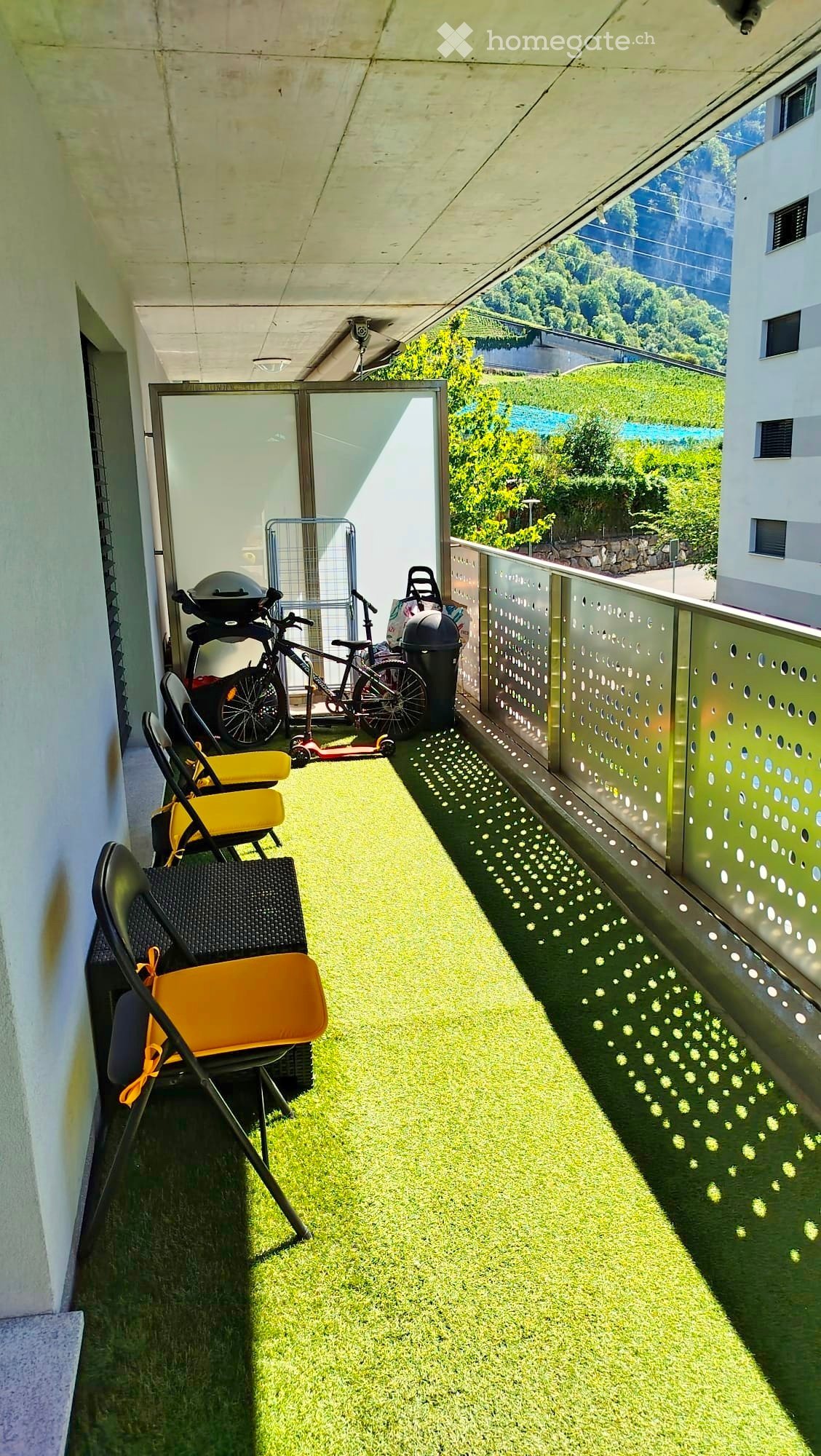 Balcony with artificial grass, chairs, bicycle, scooter, and trash bin, overlooking a scenic view.