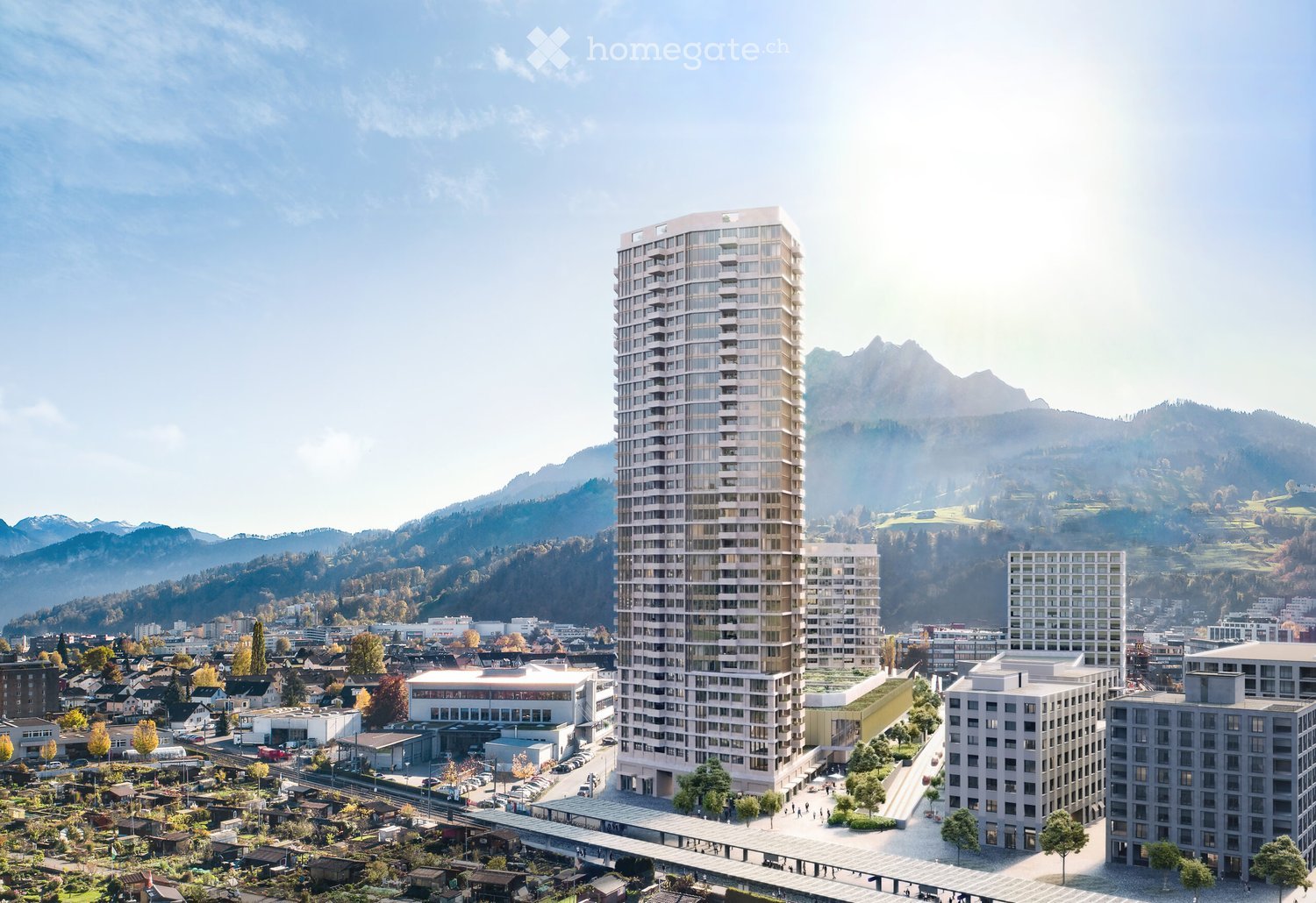 A modern high-rise building with a sleek design stands prominently in the foreground, surrounded by smaller residential structures and lush greenery. The background features rolling hills and a clear blue sky.