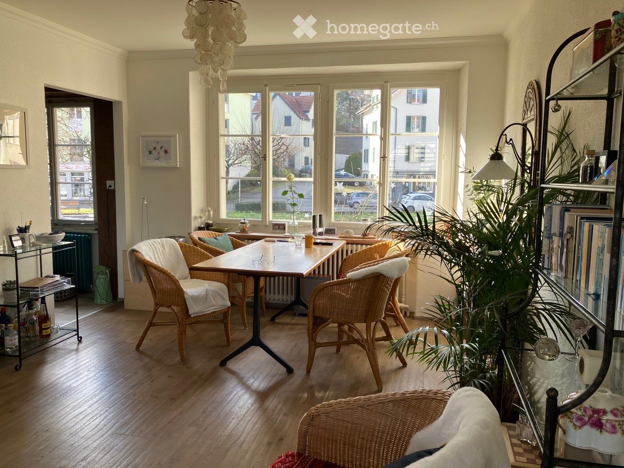 Bright living room with wooden flooring, large windows, a central table with chairs, a floor lamp, and a shelf with books and decorative items.