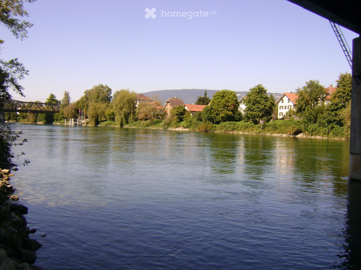 A serene river scene with calm water, a bridge in the background, and a small town with houses and trees on the opposite bank.