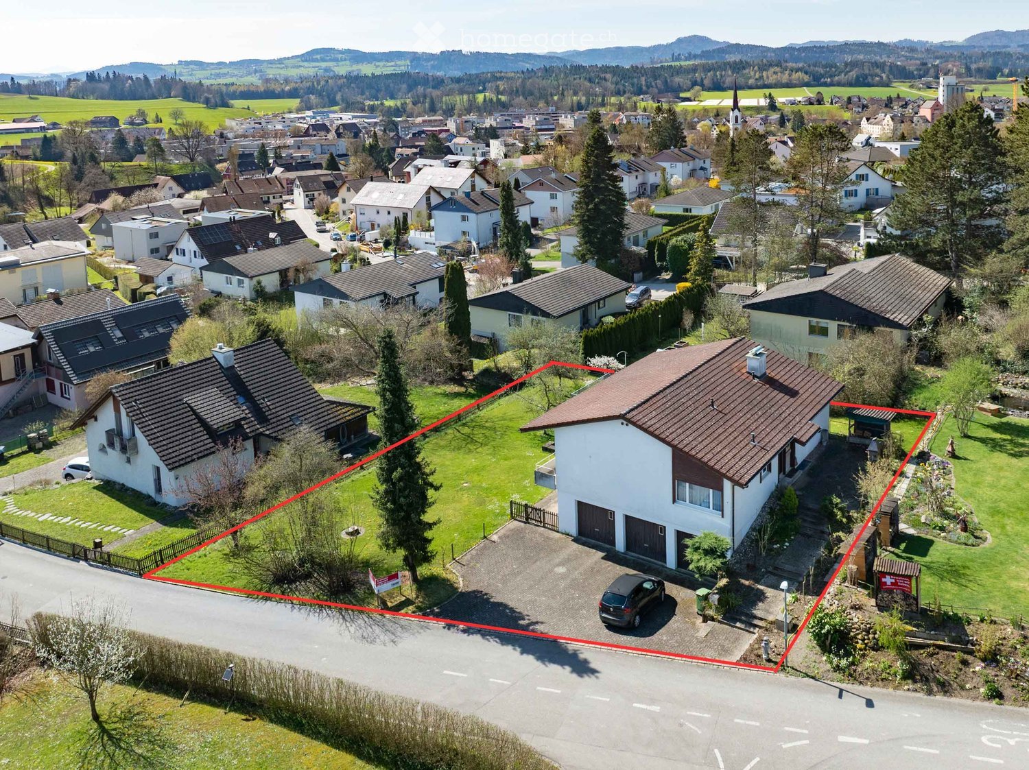Aerial view of a residential area with multiple houses, green lawns, and a driveway with a parked car. The highlighted house has a garage and a parking area.