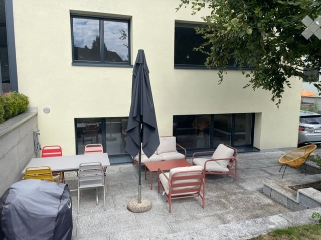 A modern patio area with a table, chairs, and a large umbrella, adjacent to a house with large windows and a sliding glass door.