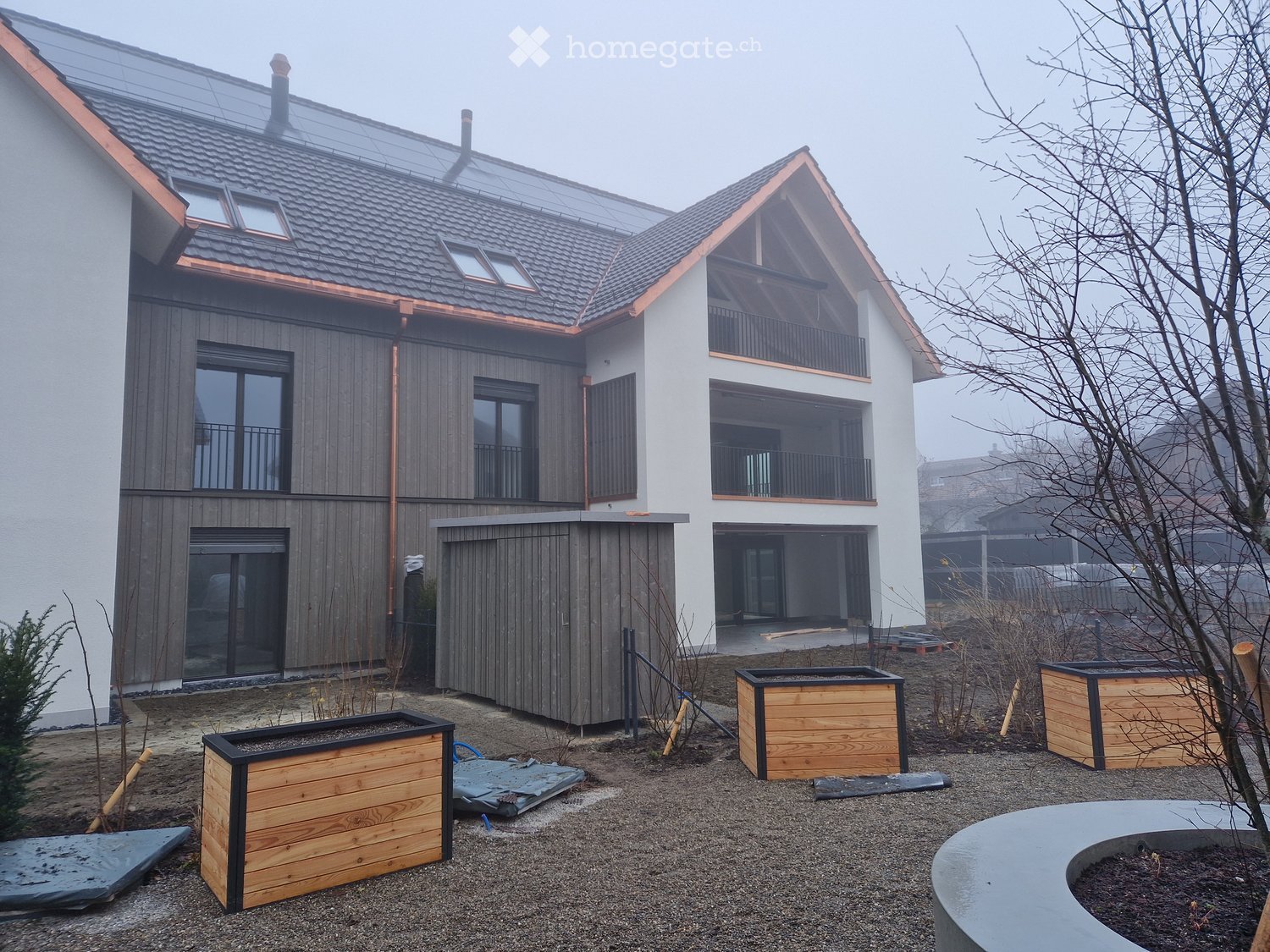 Modern two-story house with a combination of white and grey wood siding, large windows, and balconies. The roof features copper accents and skylights. The surrounding area includes wooden planters and a curved concrete bench.