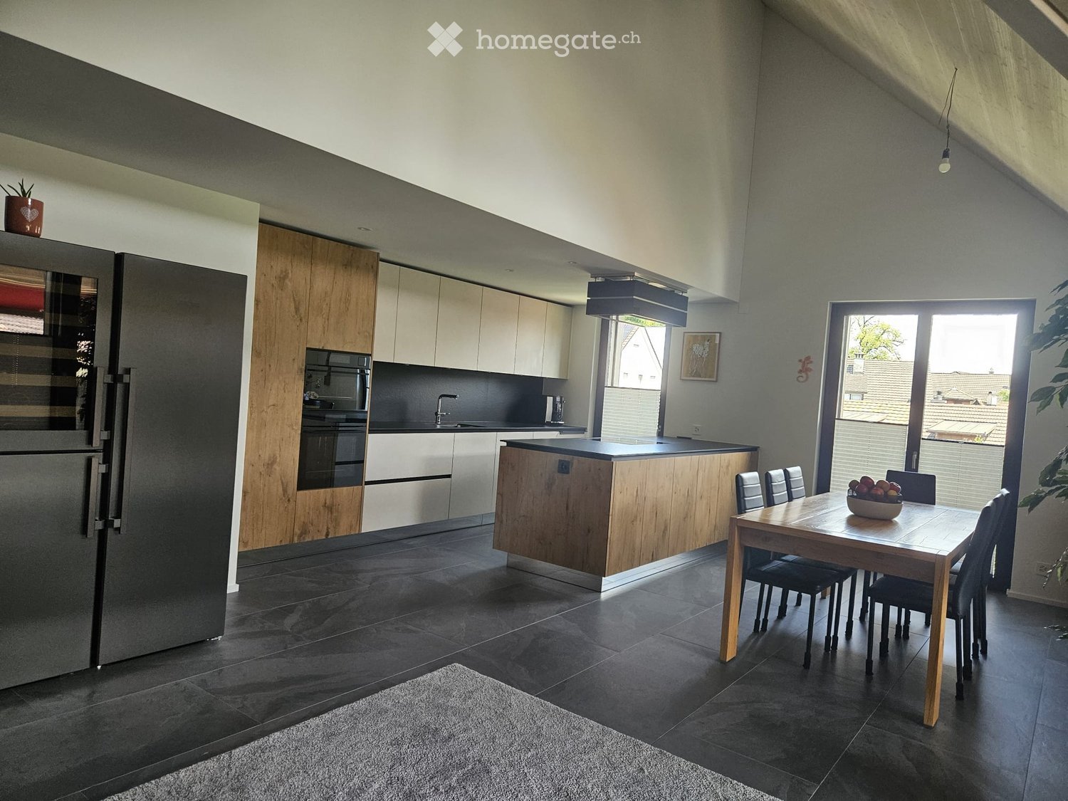 Modern kitchen with dark tiled floor, light wood cabinets, stainless steel appliances, island, and dining area with a table and chairs.