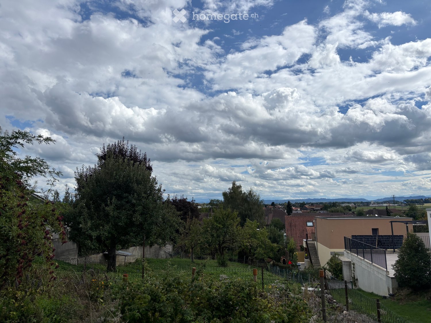 A garden area with trees, a fence, and a modern house in the background under a partly cloudy sky.