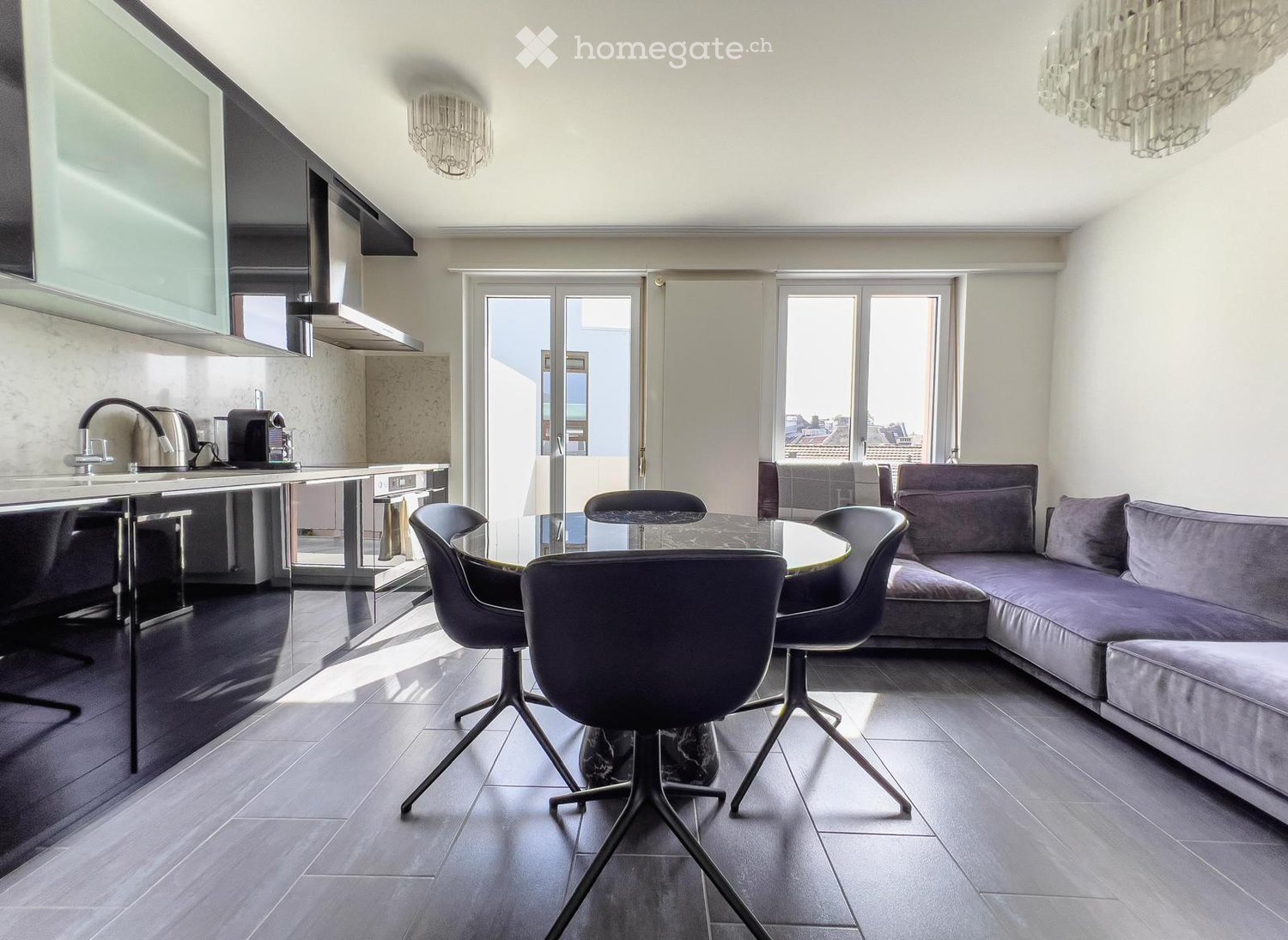 Modern open-plan living and dining area with a grey sectional sofa, glass coffee table, four chairs, and a sleek kitchen counter with appliances.