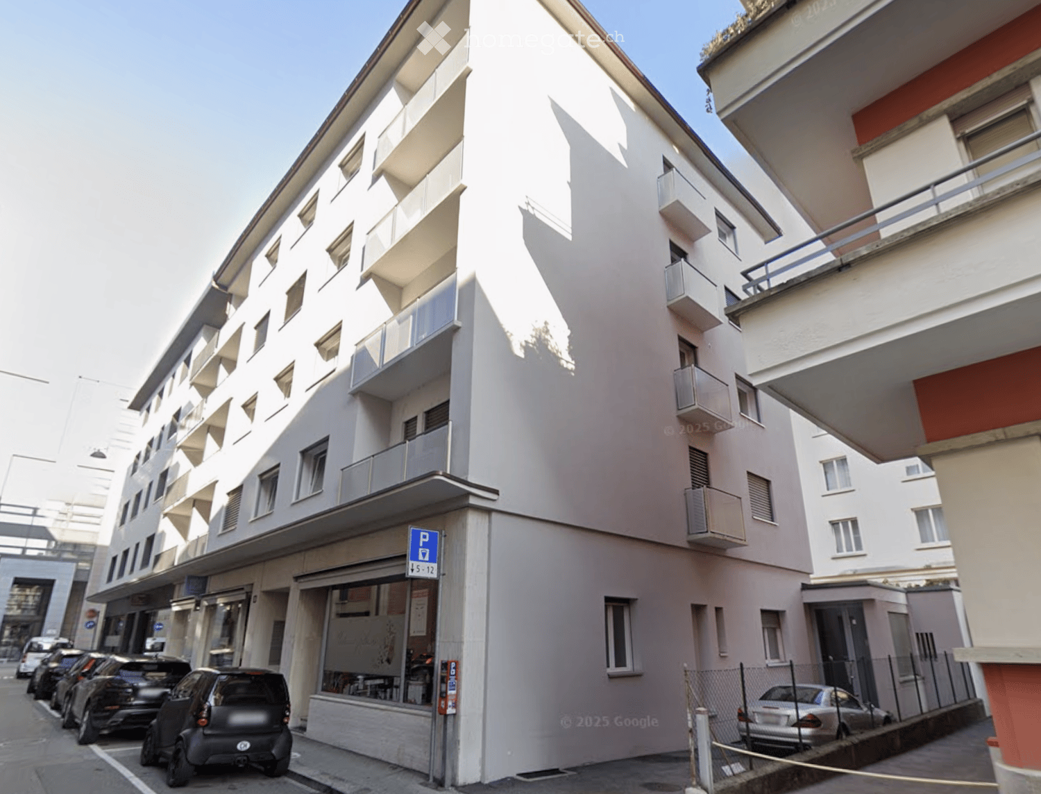 Modern multi-story building with white facade, multiple windows, and balconies. A small storefront is visible at the ground level, with several parked cars along the street.