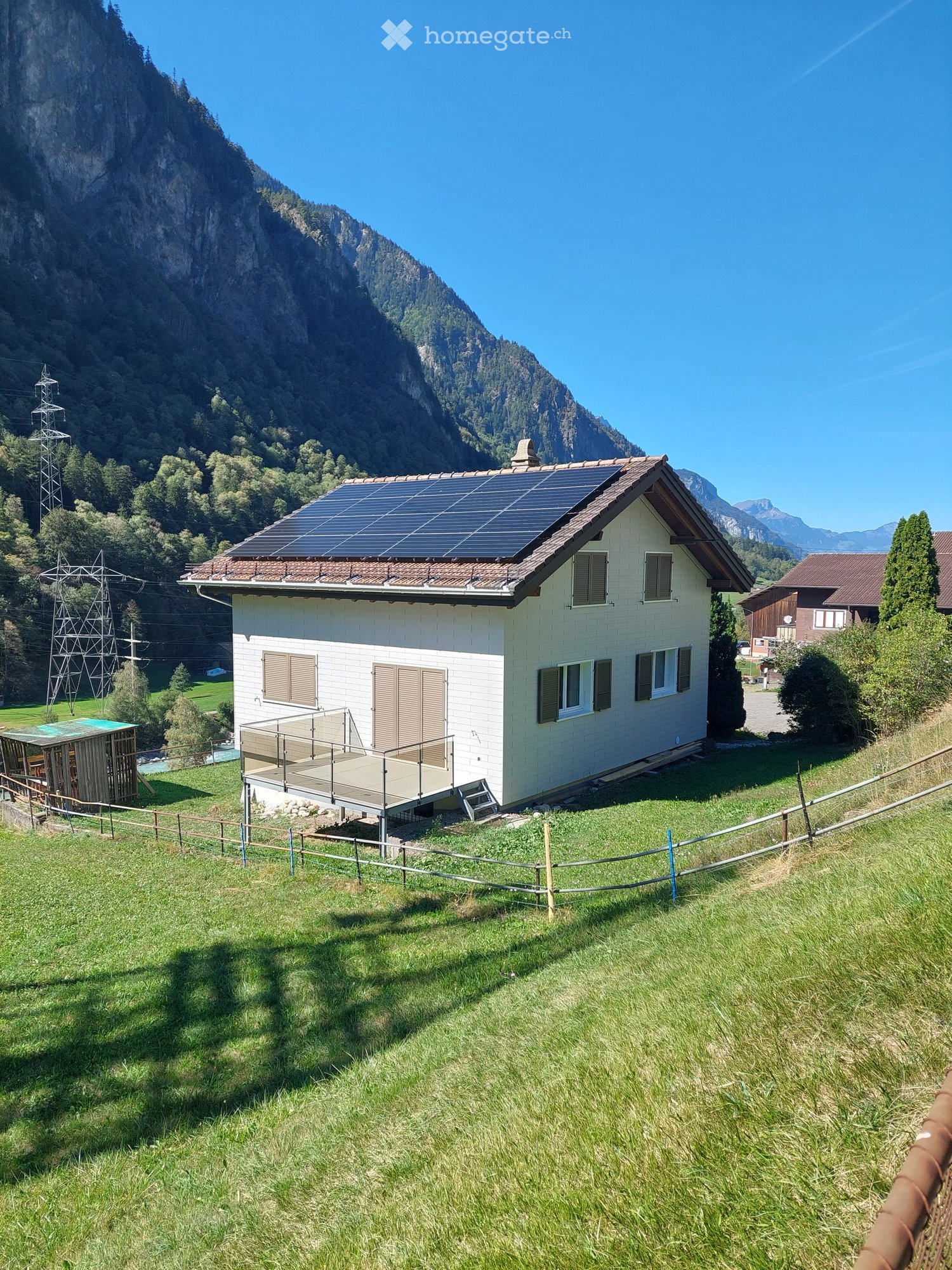 Single-story house with white walls, solar panels on the roof, and a small balcony. Surrounded by green grass and mountains.
