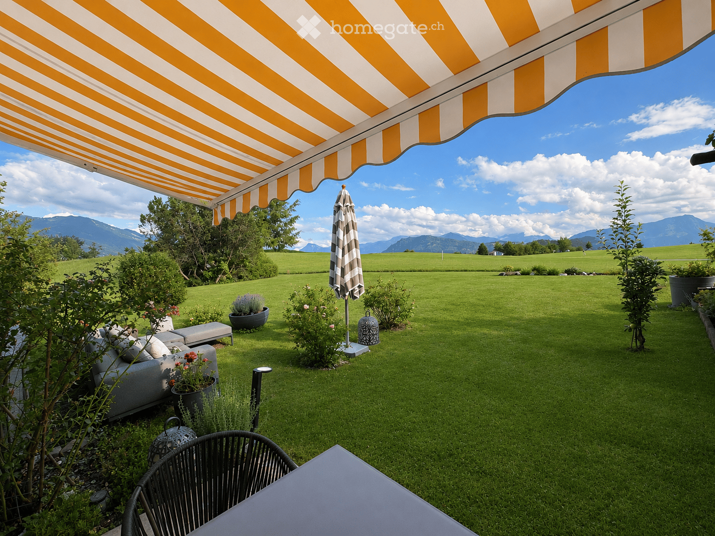 A well-maintained garden with a green lawn, a grey sofa, a black chair, a table, and a closed umbrella. The area is shaded by a yellow and white striped awning, with potted plants and a scenic mountain view in the background.