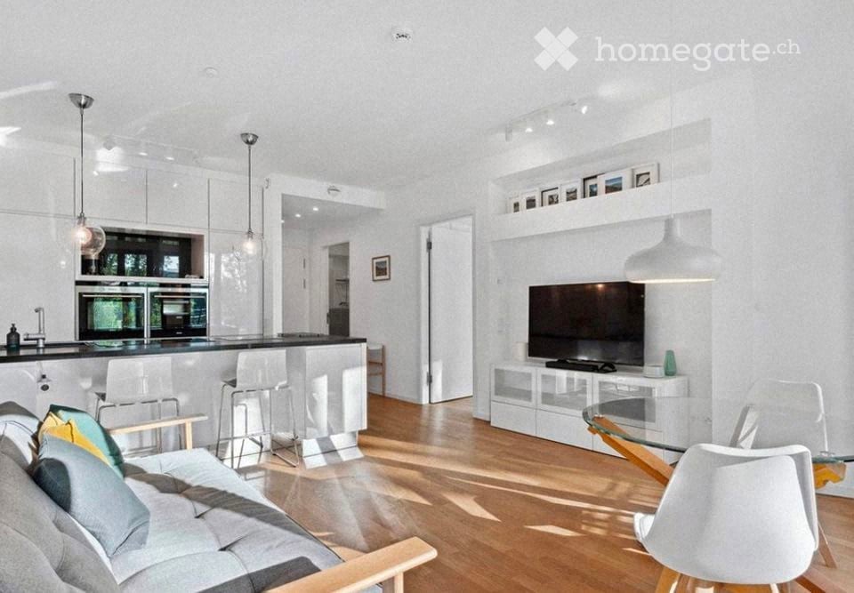Modern, open-plan living and dining area with a grey sofa, coffee table, bar stools, a kitchen island with an oven and fridge, and a television on a white stand. The space features light wooden flooring and white walls with built-in shelving.