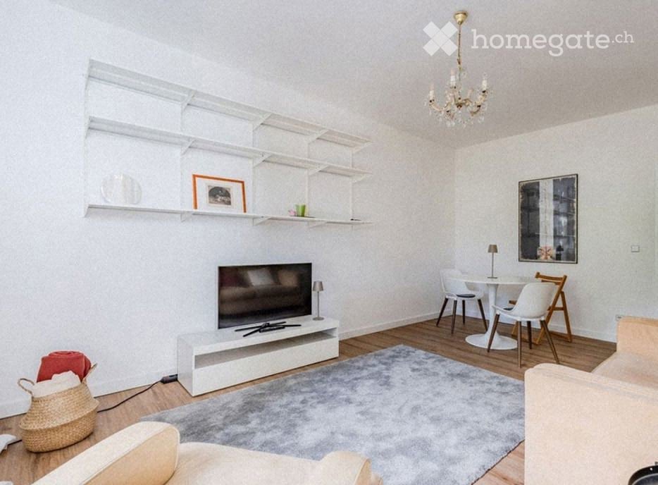 Modern living room with white walls, wooden flooring, and a grey rug. Features a white sofa, coffee table, TV stand, dining table with chairs, shelves with decorative items, and a chandelier.