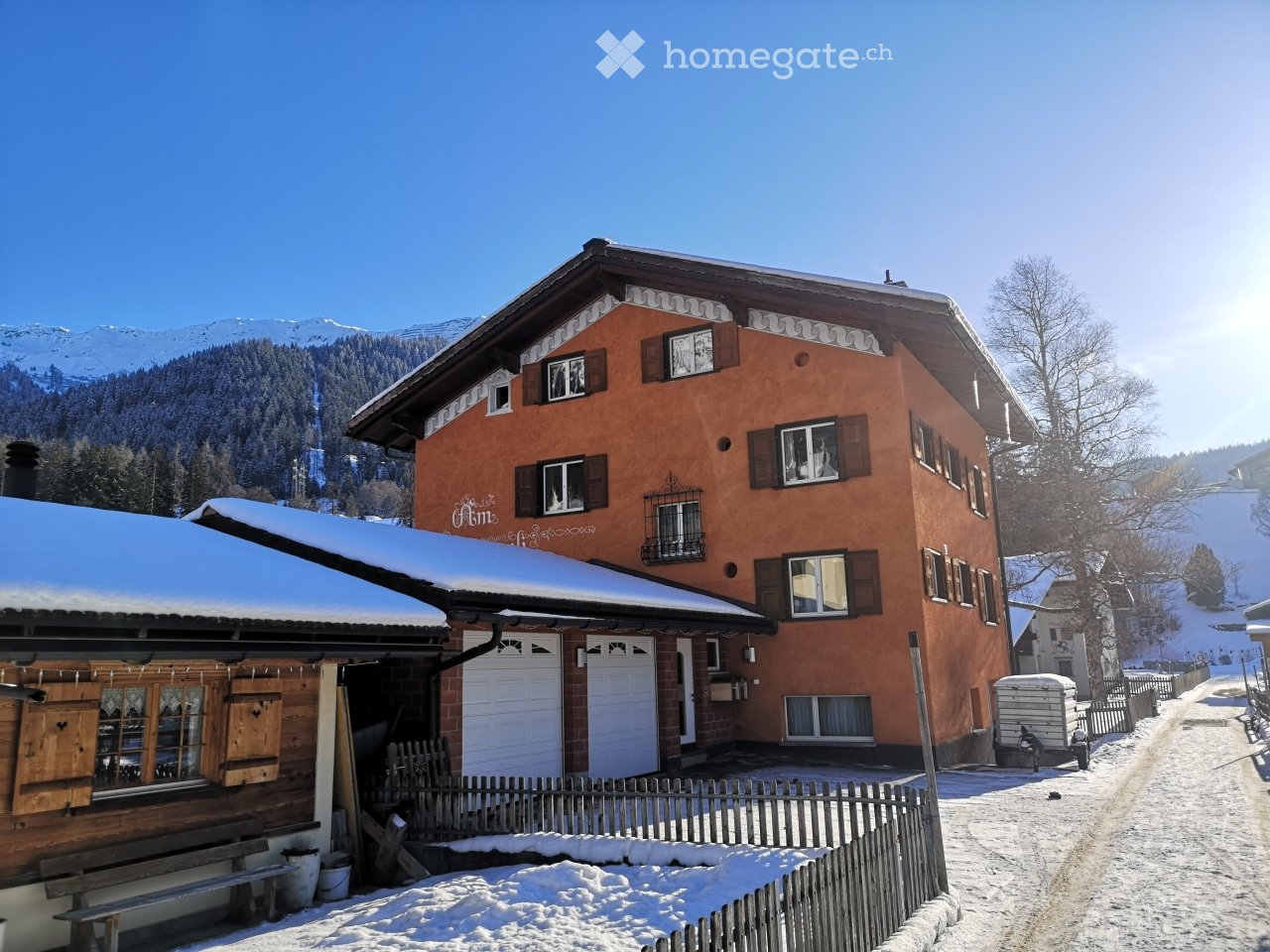 A two-story house with a red-orange facade, multiple windows, and a snow-covered roof, situated in a snowy mountain landscape.