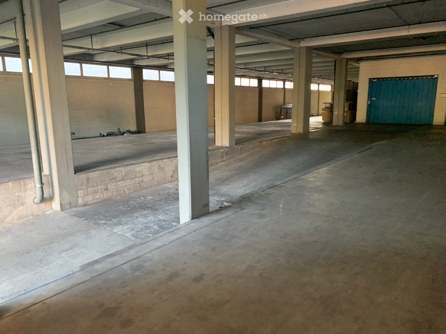 Large, empty industrial-style space with concrete floors, exposed metal beams, and minimal furnishings. Features include large windows, a raised platform area, and a green door at the far end.