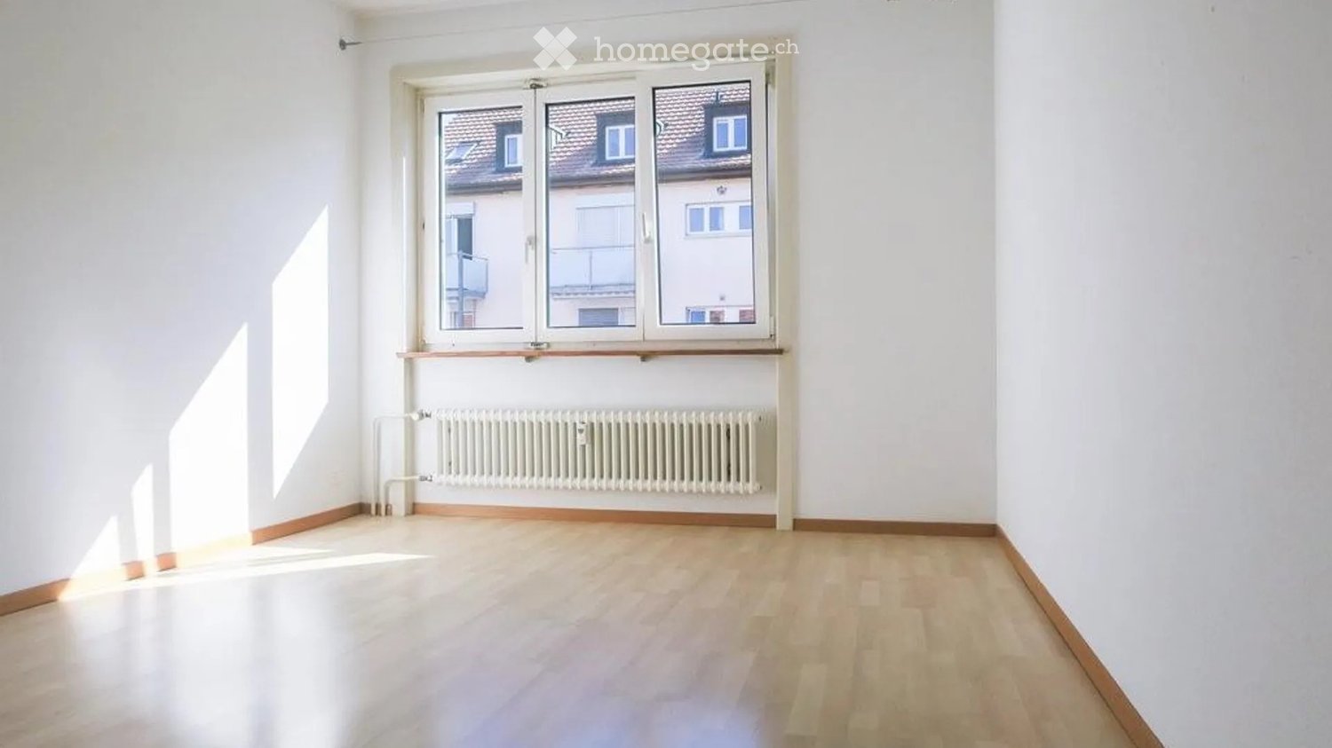 Empty room with white walls, wooden flooring, and a large window with a radiator below it.